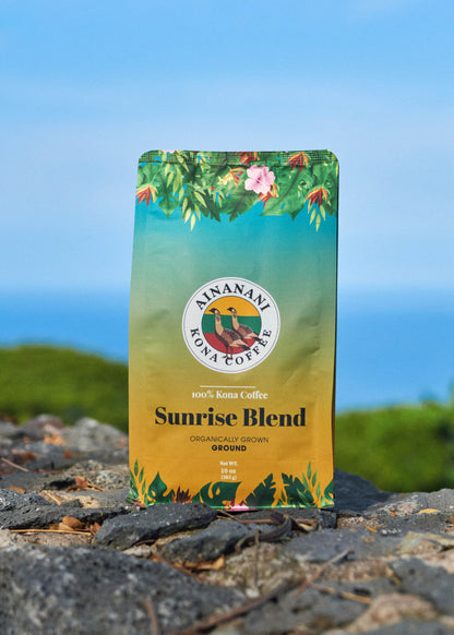 Ainanani 100% Kona Sunrise Blend ground coffee bag on lava rock above the ocean